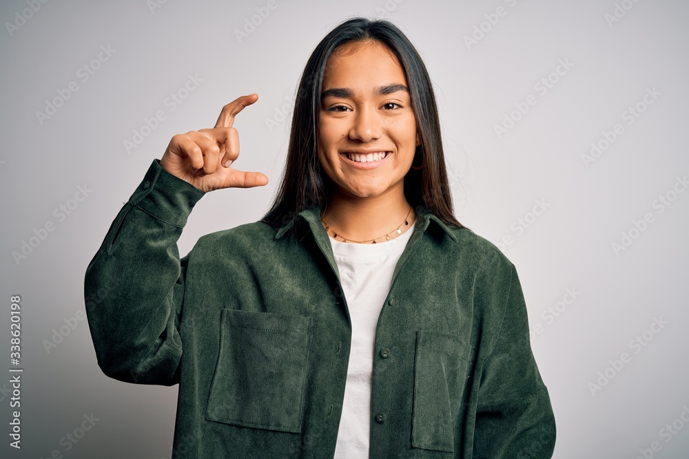 Young beautiful asian woman wearing casual shirt standing over isolated white background smiling and confident gesturing with hand doing small size sign with fingers looking and the camera. Measure