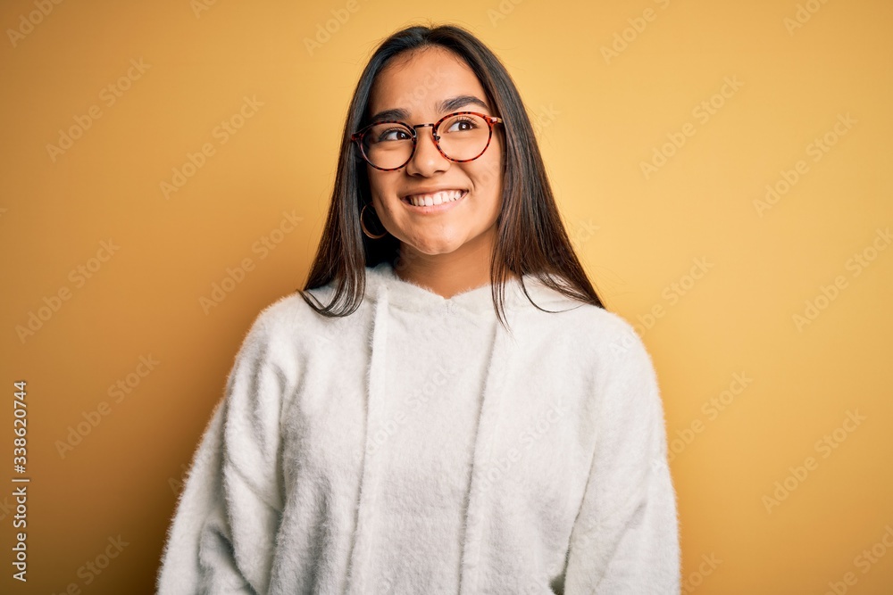 Young beautiful asian woman wearing casual sweater and glasses over yellow background looking away to side with smile on face, natural expression. Laughing confident.