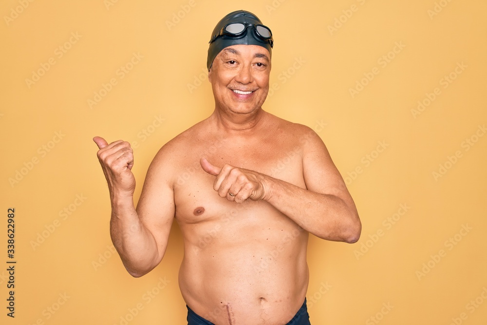 Middle age senior grey-haired swimmer man wearing swimsuit, cap and goggles Pointing to the back behind with hand and thumbs up, smiling confident