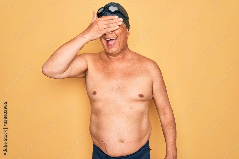 Middle age senior grey-haired swimmer man wearing swimsuit, cap and goggles smiling and laughing with hand on face covering eyes for surprise. Blind concept.