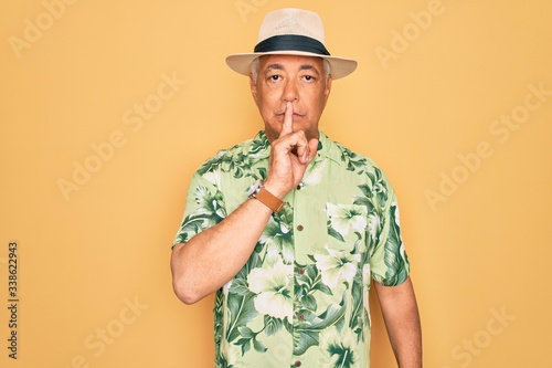 Middle age senior grey-haired man wearing summer hat and floral shirt on beach vacation asking to be quiet with finger on lips. Silence and secret concept.