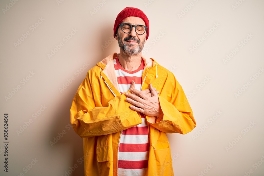 Middle age hoary man wearing glasses and rain coat standing over isolated white background smiling with hands on chest with closed eyes and grateful gesture on face. Health concept.
