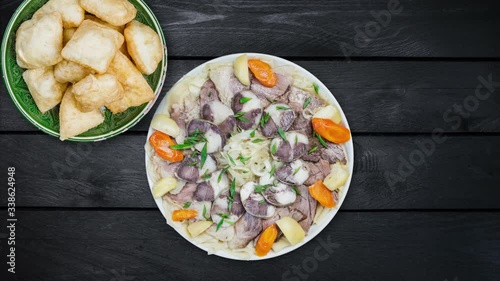 Beshbarmak dish with baursak rotates on a white plate - traditional Kazakh Asia food and national bread. Noodles with lamb and onion close-up on a plate on black wooden background. Horizontal top view photo