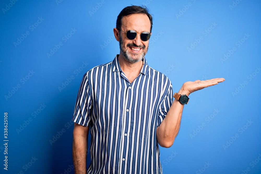 Naklejka premium Middle age hoary man wearing striped shirt and sunglasses over isolated blue background smiling cheerful presenting and pointing with palm of hand looking at the camera.