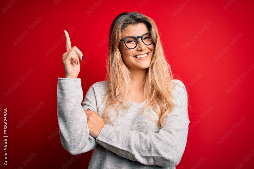 Young beautiful blonde woman wearing sweater and glasses over isolated red background with a big smile on face, pointing with hand and finger to the side looking at the camera. Young beautiful blonde woman wearing sweater and glasses over isolated red background with a big smile on face, pointing with hand and finger to the side looking at the camera.