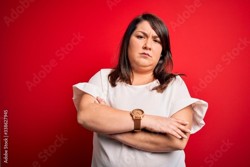 Beautiful brunette plus size woman wearing casual t-shirt over isolated red background skeptic and nervous, disapproving expression on face with crossed arms. Negative person.