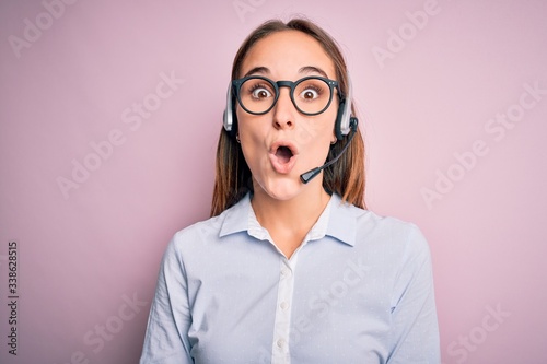 Young beautiful call center agent woman wearing glasses working using headset afraid and shocked with surprise expression, fear and excited face.