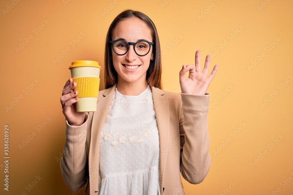 Young beautiful businesswoman drinking cup of takeaway coffee over yellow background doing ok sign with fingers, excellent symbol Young beautiful businesswoman drinking cup of takeaway coffee over yellow background doing ok sign with fingers, excellent symbol