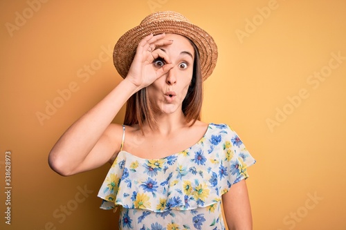 Young beautiful woman wearing casual t-shirt and summer hat over isolated yellow background doing ok gesture shocked with surprised face, eye looking through fingers. Unbelieving expression.