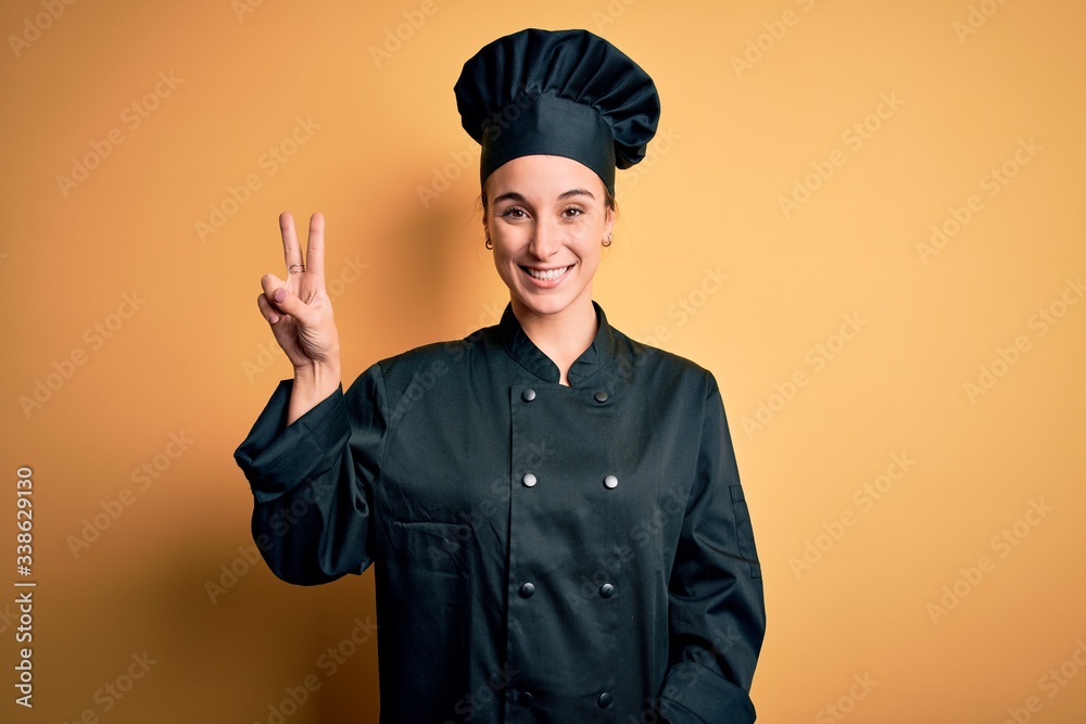 Young beautiful chef woman wearing cooker uniform and hat standing over yellow background showing and pointing up with fingers number two while smiling confident and happy. Young beautiful chef woman wearing cooker uniform and hat standing over yellow background showing and pointing up with fingers number two while smiling confident and happy.