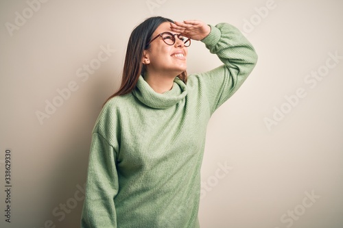 Young beautiful woman wearing casual sweater standing over isolated white background very happy and smiling looking far away with hand over head. Searching concept. © Krakenimages.com