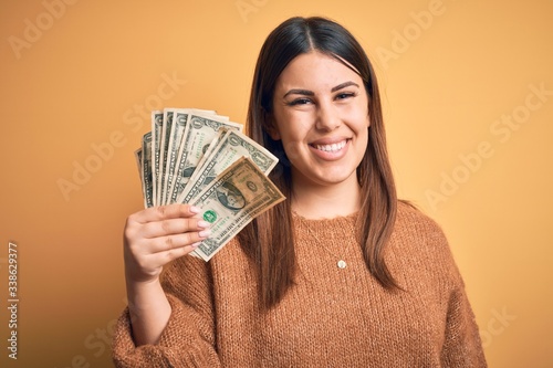 Young beautiful woman holding dollars standing over isolated orange background with a happy face standing and smiling with a confident smile showing teeth