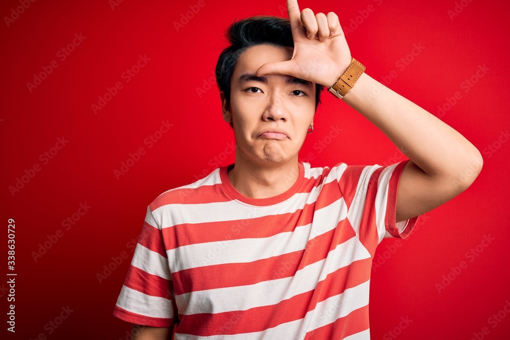 Young handsome chinese man wearing casual striped t-shirt standing over red background making fun of people with fingers on forehead doing loser gesture mocking and insulting. Young handsome chinese man wearing casual striped t-shirt standing over red background making fun of people with fingers on forehead doing loser gesture mocking and insulting.