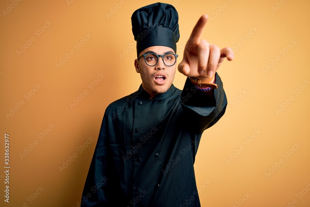 Young brazilian chef man wearing cooker uniform and hat over isolated yellow background Pointing with finger surprised ahead, open mouth amazed expression, something on the front Young brazilian chef man wearing cooker uniform and hat over isolated yellow background Pointing with finger surprised ahead, open mouth amazed expression, something on the front