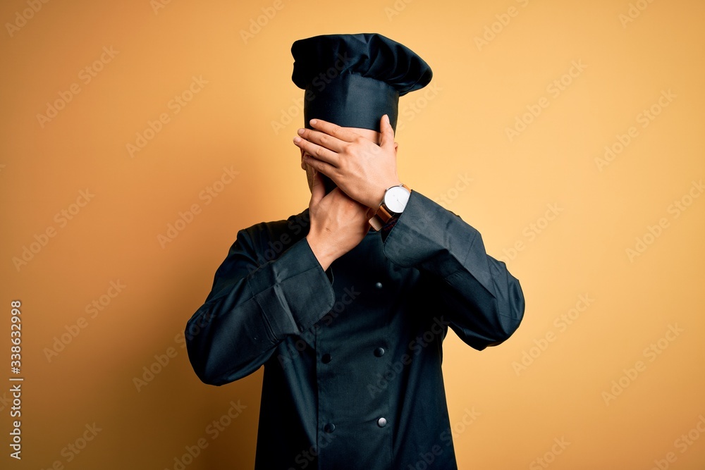 Young brazilian chef man wearing cooker uniform and hat over isolated yellow background Covering eyes and mouth with hands, surprised and shocked. Hiding emotion Young brazilian chef man wearing cooker uniform and hat over isolated yellow background Covering eyes and mouth with hands, surprised and shocked. Hiding emotion