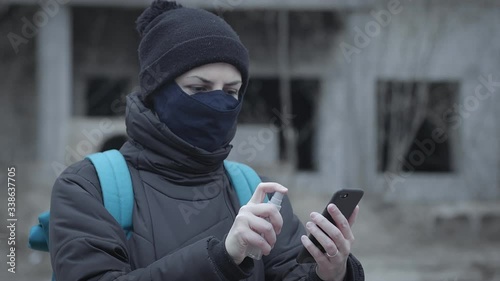 Young woman in a protective mask sprays antiseptic on her smartphone against the background of an abandoned building in an industrial post-apocalyptic landscape. Pandemic protection, desinfection