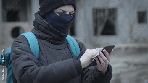 Young woman in a protective mask cleans her smartphone with sanitary napkin against the background of an abandoned building in an industrial post-apocalyptic landscape. Pandemic protection