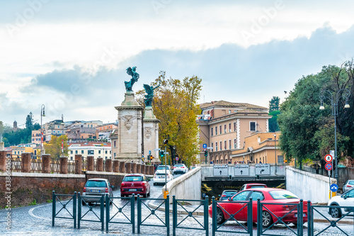 Fototapeta Naklejka Na Ścianę i Meble -  Rome, Italy. Vehicular traffic on cobblestone streets of the city. Statues of angels with wreaths and sword, underground road/ roadway, Roman architecture buildings in background.