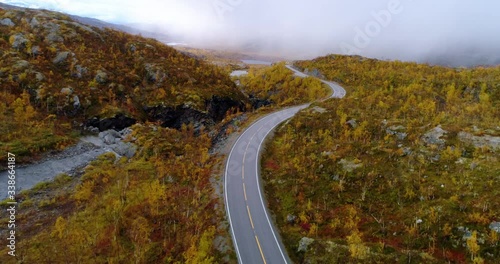 Mountain road in foliage forest, Aerial, rising, drone shot, overlooking a asphalt route in arctic wilderness, rain clouds in background, dark, fall day, in Skibotn, Nordkalotten, North Norway photo