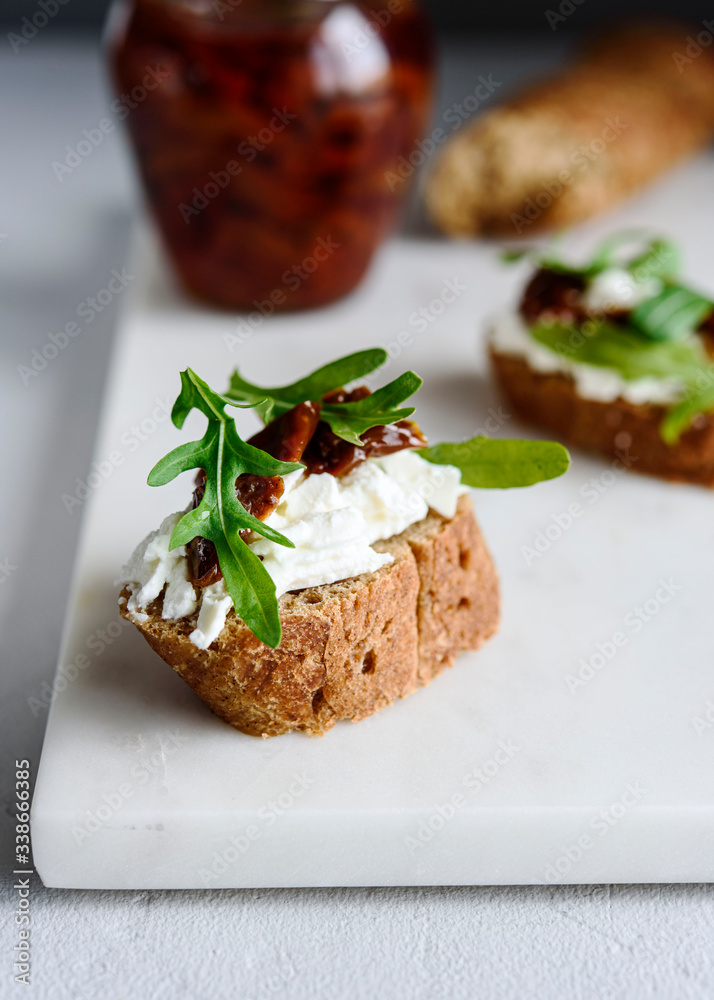 Italian bruschetta (sandwich, crostini, toast) with confit tomatoes, feta, olive oil and arugula leaves on white marble background. Traditional italian appetizer or snack, antipasto. Selective focus