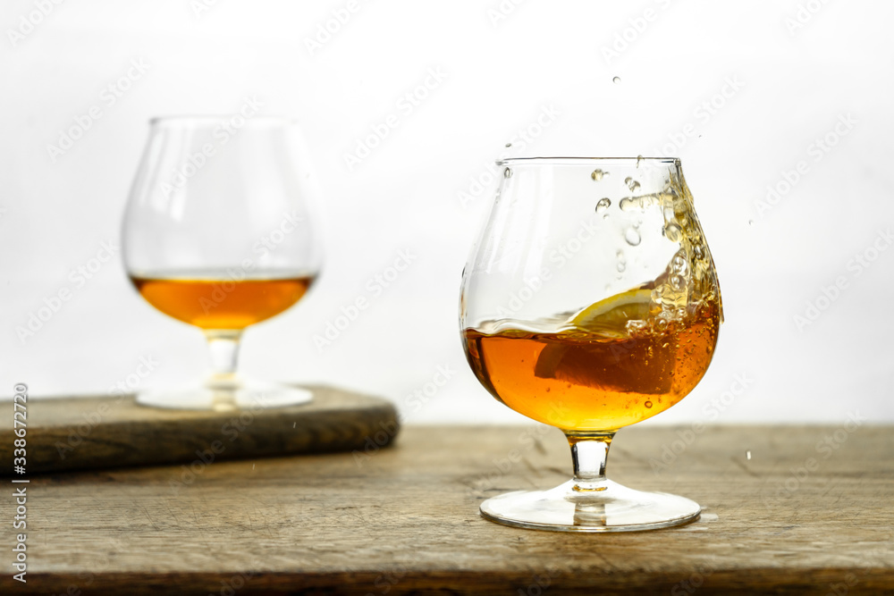 Cognac \ whiskey in cognac glass on wooden table, golden color spirits splashing on white background Cognac \ whiskey in cognac glass on wooden table, golden color spirits splashing on white background
