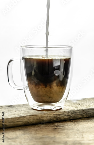 Pouring milk into the black coffee in transparent cup on wooden table