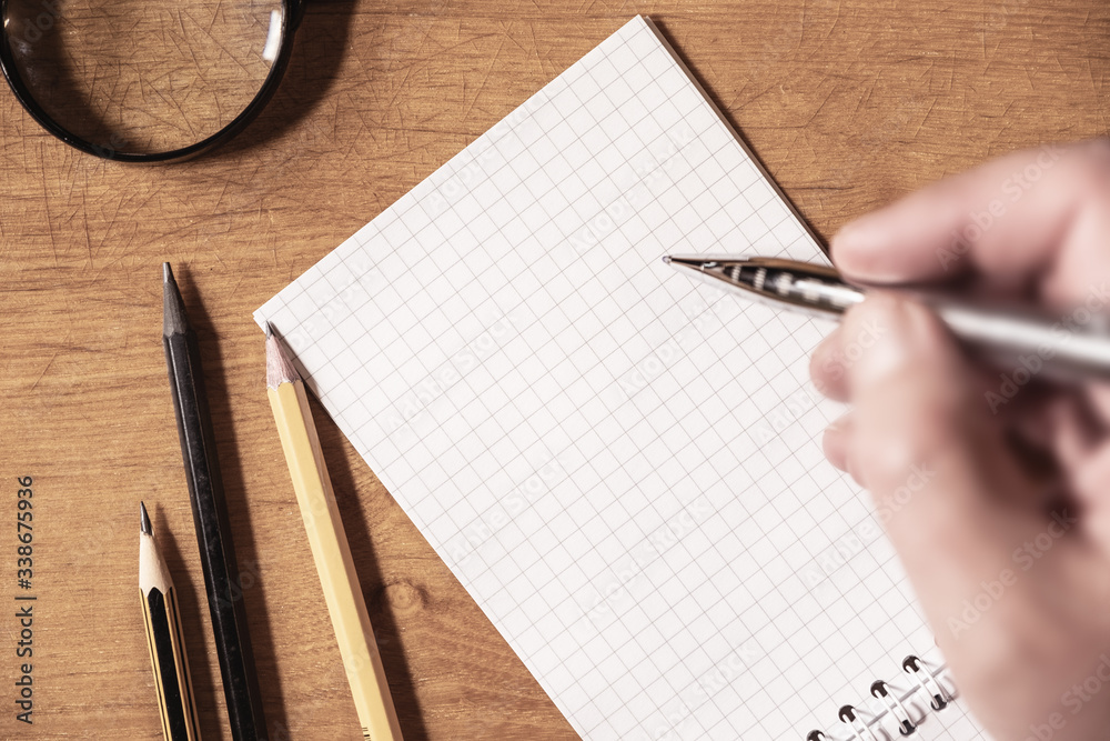 Top view of man's hand writing on a blank paper. Pencils and magnifier on the table. Top view of man's hand writing on a blank paper. Pencils and magnifier on the table.