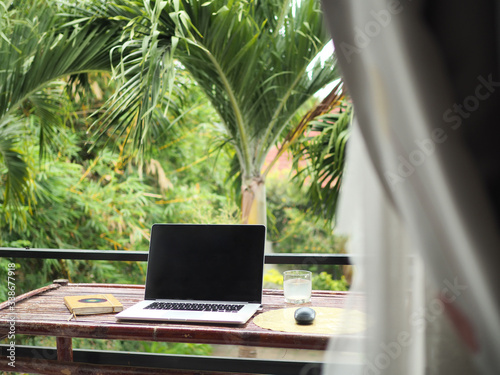 laptop of a remote digital nomad on a wooden bamboo table with notebook, mobile phone and glass in nature on a balcony with a green tropical background with palm trees photo