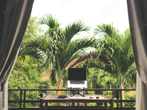 laptop of a remote digital nomad on a wooden bamboo table with notebook, mobile phone, glass and chair in nature on a balcony with a green tropical background with palm trees photo