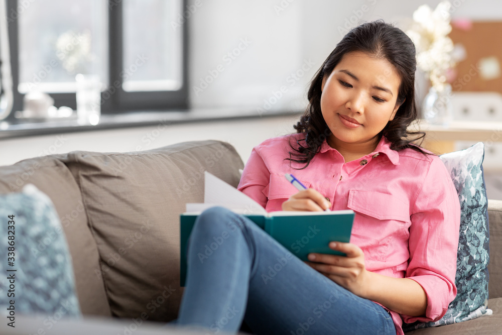 people and leisure concept - happy smiling asian young woman with diary or notebook sitting on sofa at home people and leisure concept - happy smiling asian young woman with diary or notebook sitting on sofa at home