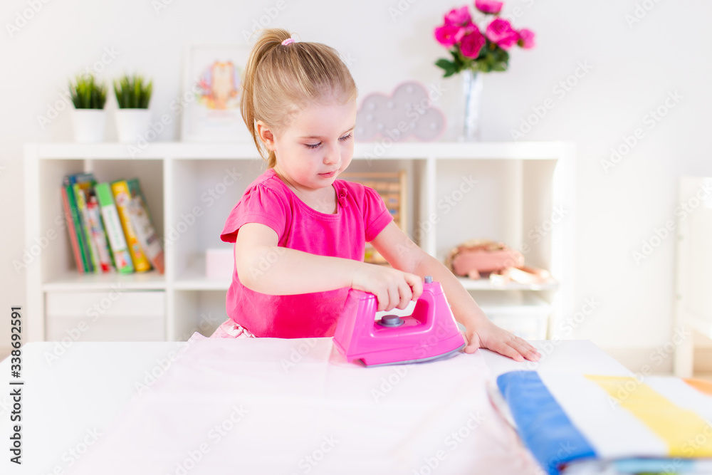 Little blonde baby girl housewife playing with toy pink iron. Ironong clothes on a white table. Housework, helper Little blonde baby girl housewife playing with toy pink iron. Ironong clothes on a white table. Housework, helper