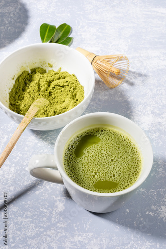Matcha green tea in white cup, matcha green tea powder, special wooden spoon, bamboo whisk, fresh leaves on grey textured backdrop with shadows. Natural organic product with antioxidants for healthy.
