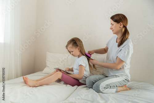 Happy young mother with daughter in the light bedroom. The daughter is engaged on the tablet while mom is combing her hair. Family relationships and home leisure. Good mood