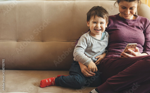 Front view portrait of a caucasian mother and son sitting on the sofa and using a mobile while smiling photo