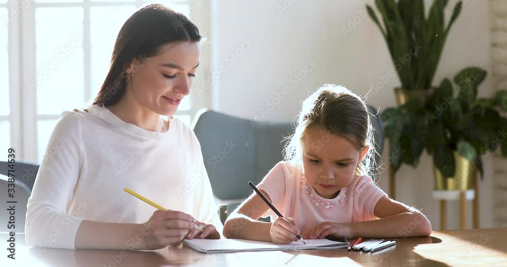 Head shot young nanny babysitter sitting at table with happy adorable small kid girl, drawing pictures in paper album with colorful pencils. Smiling mommy enjoying hobby weekend time with daughter. Head shot young nanny babysitter sitting at table with happy adorable small kid girl, drawing pictures in paper album with colorful pencils. Smiling mommy enjoying hobby weekend time with daughter.