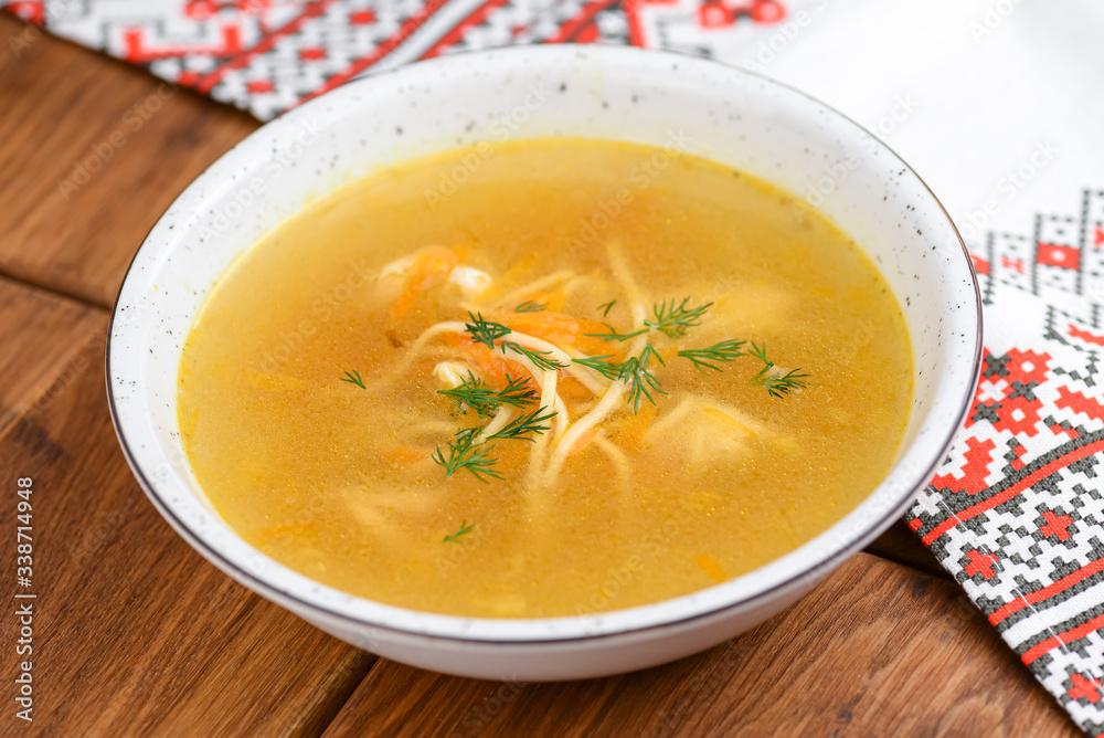 Chicken soup with noodles and vegetables served in white bowl over rustic wooden table with traditional tablecloth. Chicken soup with noodles and vegetables served in white bowl over rustic wooden table with traditional tablecloth.