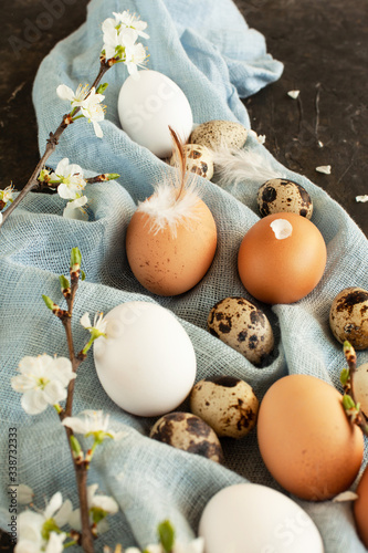 Beautiful Easter eggs card. Mix of white and brown chicken and quail eggs of natural color on blue linen towel with feathers and small white tree flowers, close up. Easter still life, selected focus.