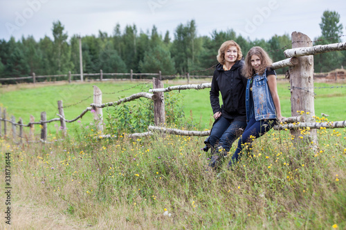 Mature mother and teenage daughter portrait on countryside, sitting together on wooden fence in pasture, female farmers, copyspace