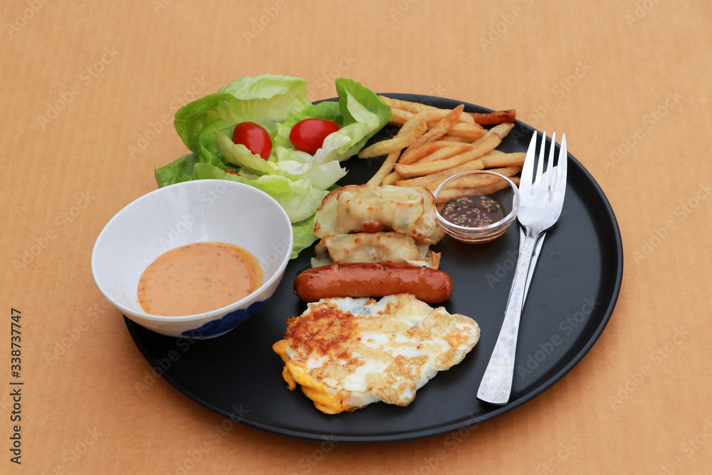 Breakfast in the black round plate on the brown floor. Fried egg with sausage, dumpling with french fries and vegetable with tomato.