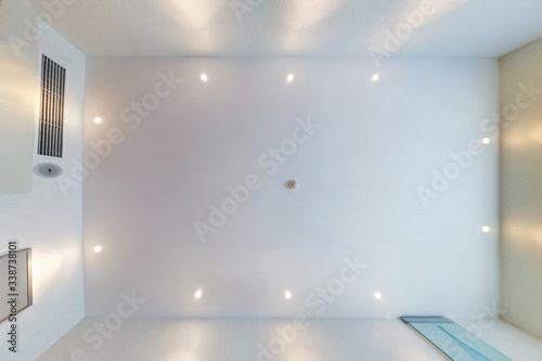 look up on suspended ceiling with halogen spots lamps and drywall construction with fire alarm sensor in empty room in apartment or house. Stretch ceiling white and complex shape.