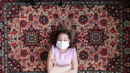 Young kid wearing protective mask. Teenager girl lying on red carpet. Quarantine, self isolation, depression, social distance, mental health concept. Stock time lapse video. photo