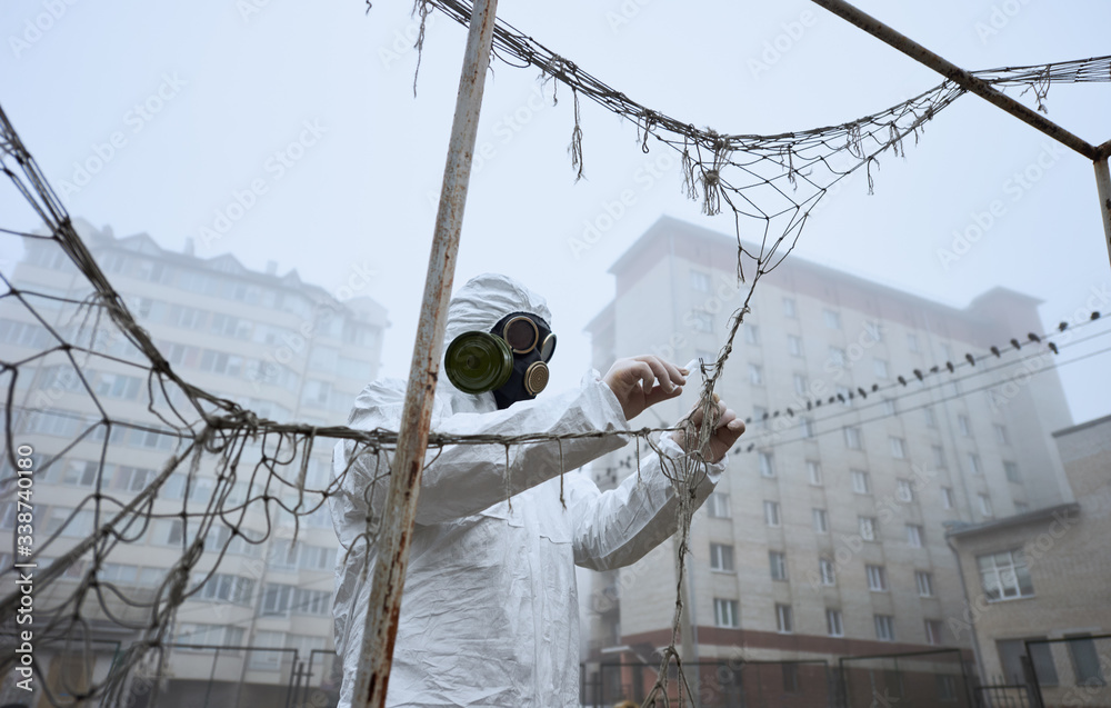 From below view of male scientist in gas mask and protective clothing monitoring abandoned football pitch, fog, buildings around. Man taking piece of goal net using tweezers and putting in test tube. - obrazy, fototapety, plakaty 