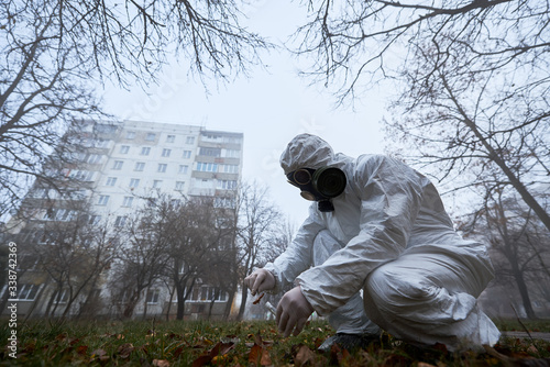 Man in gas mask and protective clothing monitoring city. From below view of scientist checking fallen tree leaves on ground, taking piece using tweezers for analysis and putting in test tube.
