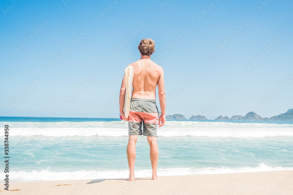 Attractive surfer man standing with surfboard under arms on the sandy beach checking the waves. Sports travel destination. Surfing lifestyle.