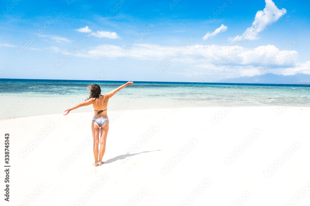Happy attractive woman in bikinis standing with extended arms on an empty wild tropical sandy beach on a sunny summer day.