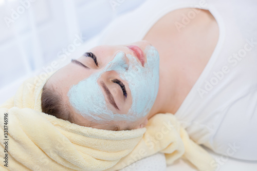 Portrait of beautiful woman laying with towel on the head. Young girl enjoys cream facial mask. Lady getting spa treatment at beauty salon. Cosmetology and skincare concept.