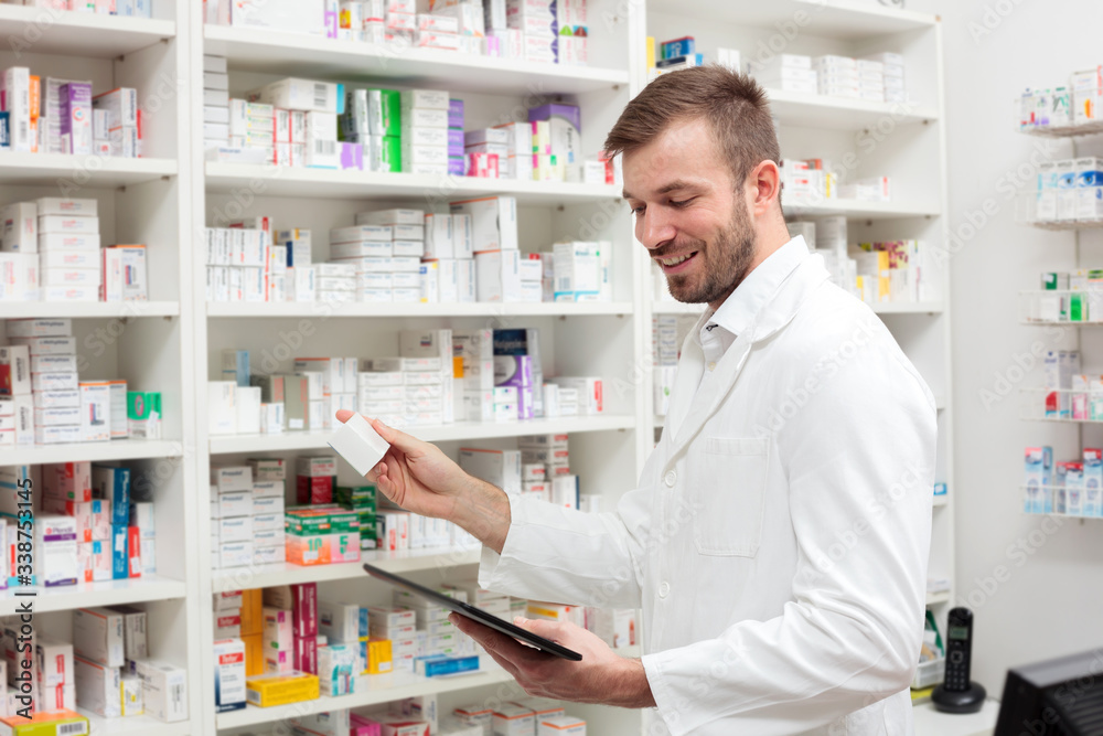 Happy young male pharmacist working in the pharmacy, holding a box of medications and using a tablet. Healthcare and medicine concept.