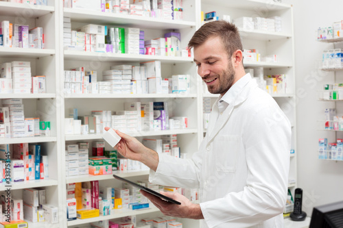 Happy young male pharmacist working in the pharmacy  holding a box of medications and using a tablet. Healthcare and medicine concept.