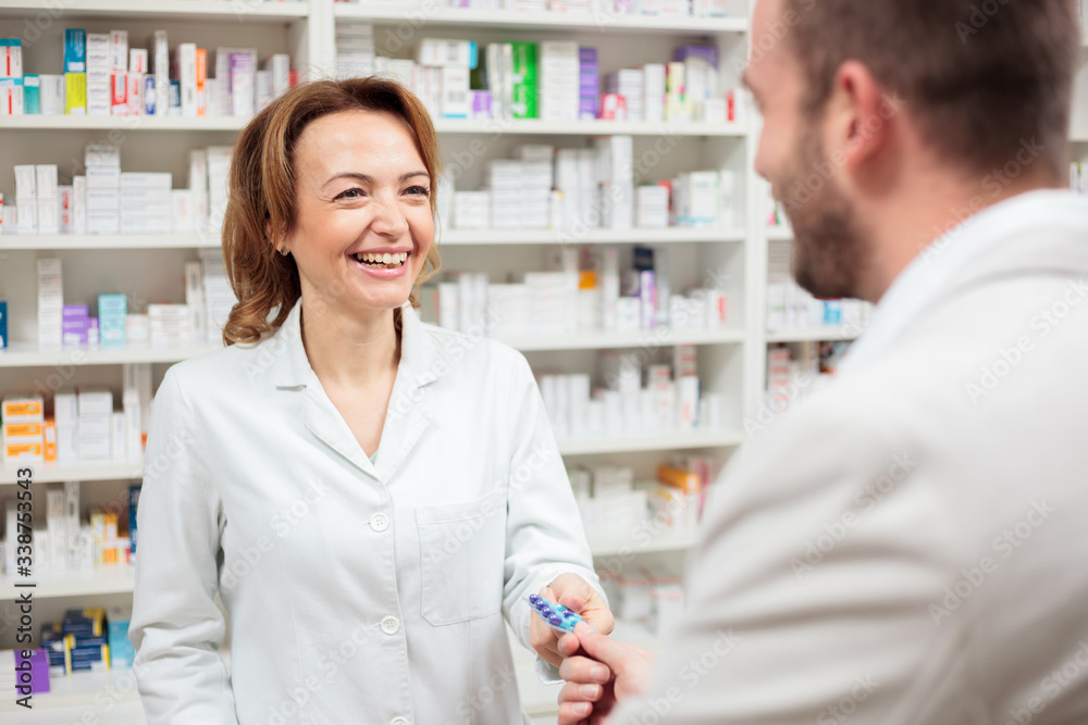 Smiling happy middle aged female pharmacist standing behind the counter, selling and giving pills to a young man. Healthcare and medicine concept.