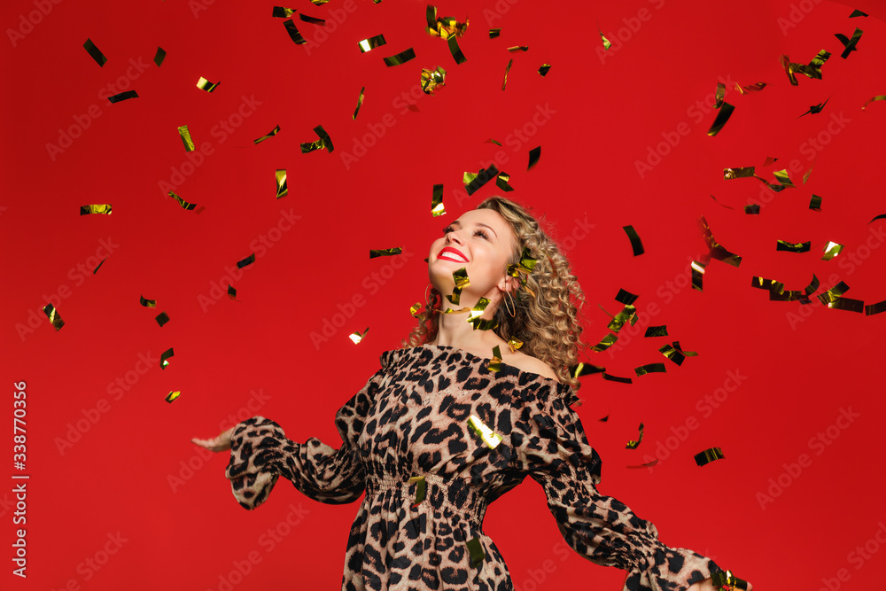 Obraz premium Portrait of a cheerful beautiful girl with curl hair standing under confetti rain and celebrating isolated over red background - Image
